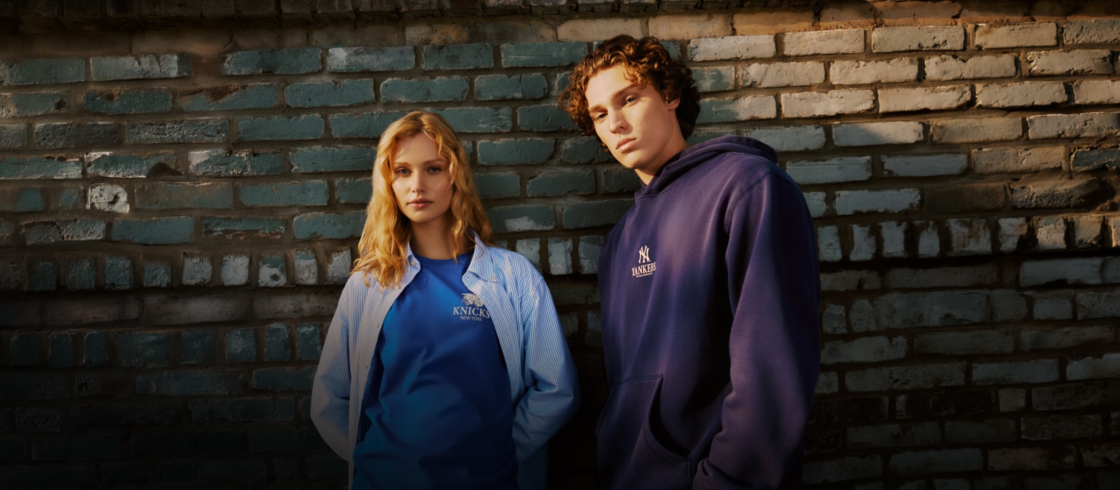 Two models wearing Mitchell & Ness Knicks and Yankees core essentials against a painted brick wall.